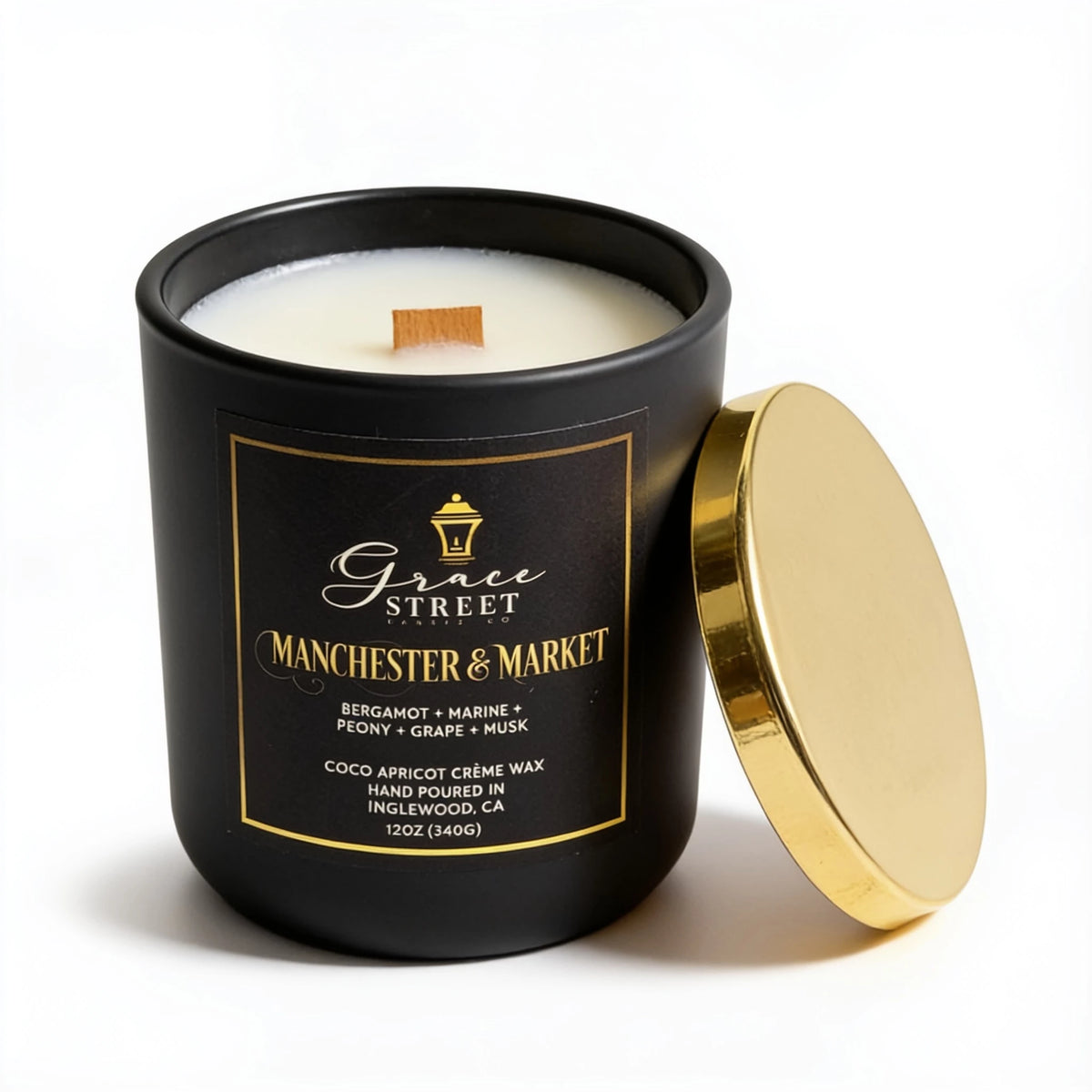 A black glass jar candle with cream-colored wax and a wooden wick, featuring a black label with gold lettering that reads Grace Street Manchester & Market. The gold metal lid is propped against the right side of the jar on a clean white background.