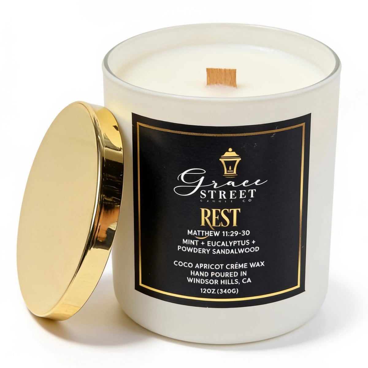 A white glass jar candle with a gold lid leaning against it on a white background. The front label is black with gold text reading Grace Street Rest.